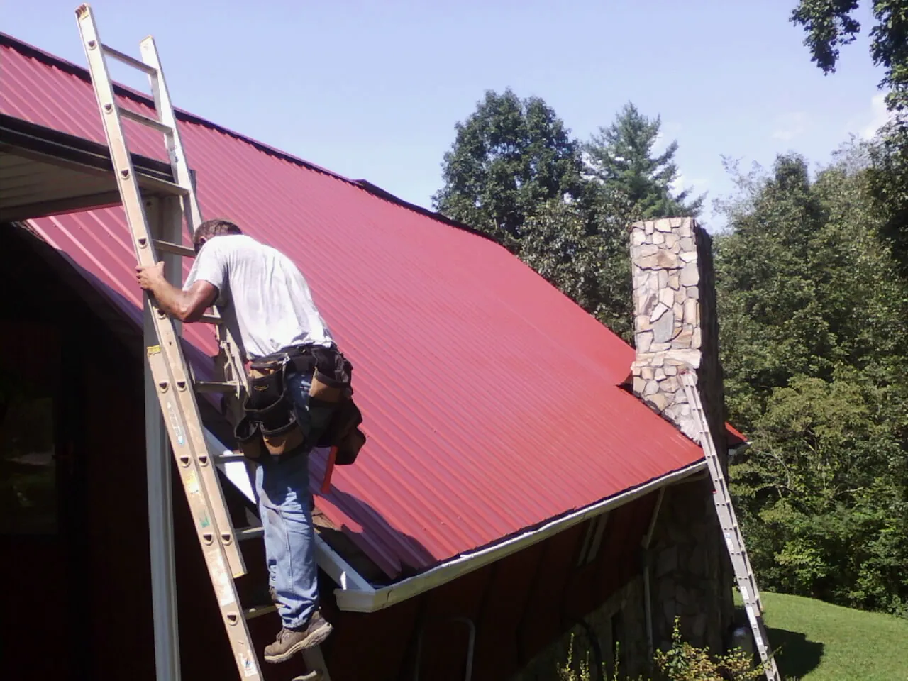 Roofer on a ladder beside a red metal roof and stone chimney — Soffit & Fascia Repair by Roofing Brothers in Staplehurst
